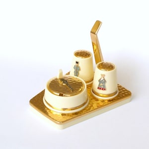 May include: A gold-toned metal salt and pepper shaker set with a matching tray. The shakers are shaped like thimbles and feature small, colorful figures. The tray has a hammered texture.