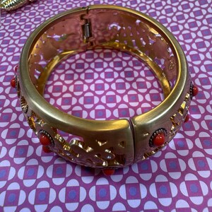 May include: Gold-tone hinged bracelet with openwork floral design and red accents. The bracelet features a clasp closure and is adorned with small, round red stones. A second bracelet is partially visible in the background.
