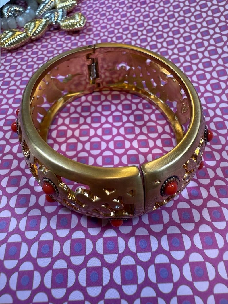 May include: Gold-tone hinged bracelet with openwork floral design and red accents. The bracelet features a clasp closure and is adorned with small, round red stones. A second bracelet is partially visible in the background.