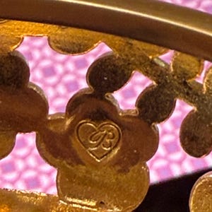 May include: Close-up of a gold-toned metal decorative piece with a floral design. The piece features a heart-shaped emblem with the initials "B" and "R" intertwined. The background is a pink patterned fabric.
