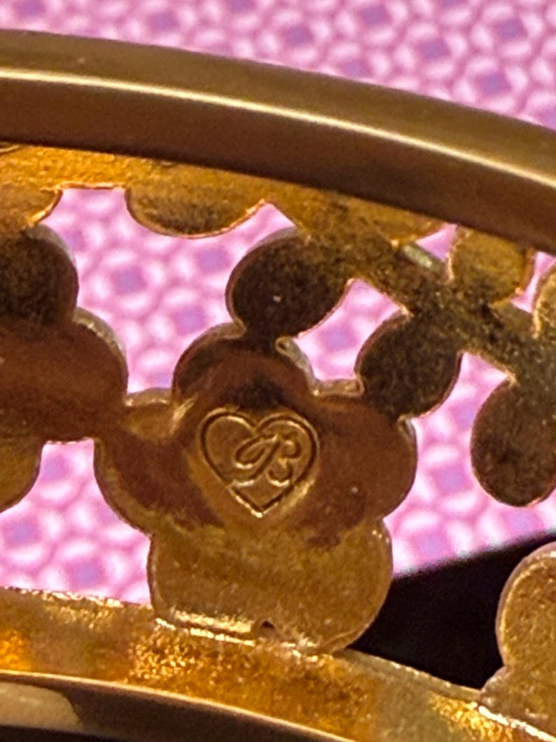 May include: Close-up of a gold-toned metal decorative piece with a floral design. The piece features a heart-shaped emblem with the initials "B" and "R" intertwined. The background is a pink patterned fabric.