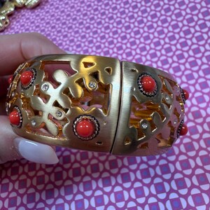 May include: Gold-tone hinged bracelet with a floral cutout design. The bracelet features red cabochon accents and small clear crystals. The bracelet is shown against a patterned pink background.