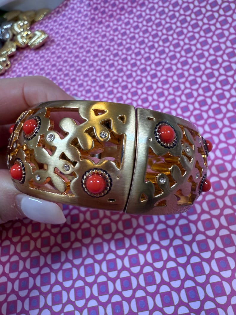 May include: Gold-tone hinged bracelet with a floral cutout design. The bracelet features red cabochon accents and small clear crystals. The bracelet is shown against a patterned pink background.