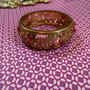 May include: Gold-tone metal bracelet with a floral cutout design. The bracelet features small, round red accents. The bracelet is shown on a patterned pink and purple surface. Other gold-tone jewelry pieces are visible in the background.