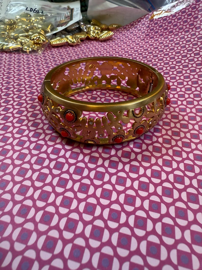 May include: Gold-tone metal bracelet with a floral cutout design. The bracelet features small, round red accents. The bracelet is shown on a patterned pink and purple surface. Other gold-tone jewelry pieces are visible in the background.