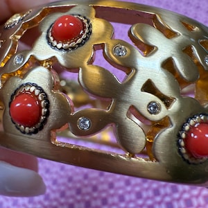 May include: Gold-toned bracelet with a floral design. The bracelet features red cabochon stones and small clear crystals. The openwork design allows light to pass through, creating a delicate and elegant look. A statement piece of jewelry.