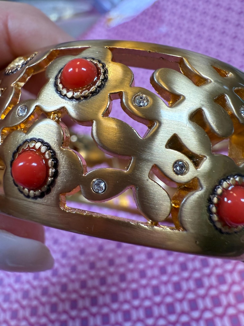 May include: Gold-toned bracelet with a floral design. The bracelet features red cabochon stones and small clear crystals. The openwork design allows light to pass through, creating a delicate and elegant look. A statement piece of jewelry.