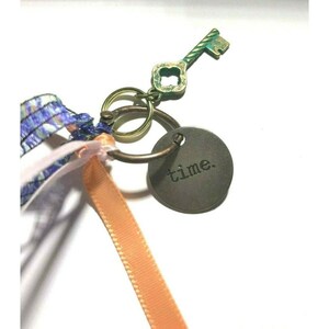 May include: A key charm with a green and gold key, a round metal tag with the word "time" engraved on it, and three ribbons in pink, blue, and white.