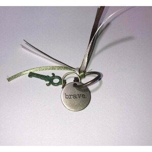 May include: A silver key chain with a small key and a round charm that says "brave." The charm is attached to a ring with a green ribbon.
