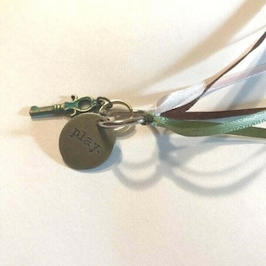 May include: A silver key charm with a green patina and a round tag that says "play." The charm is attached to a brown, white, and green ribbon.