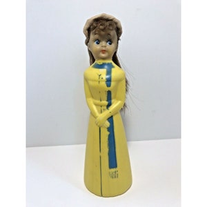 May include: A yellow plastic doll wearing a brown hat and a blue scarf. The doll has brown hair, blue eyes, and a painted face.