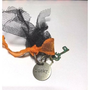 May include: A silver charm with the word "lucky" engraved on it, attached to a key and a black tulle bow with an orange ribbon.