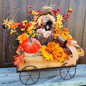 May include: A rustic wooden wagon filled with fall foliage, including orange and yellow leaves, a small scarecrow doll, and a velvet pumpkin. The wagon has black metal wheels and a gingham ribbon.
