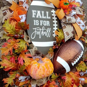 May include: A fall-themed wreath with a brown football-shaped sign that says "Fall is for Football". The wreath is decorated with orange and brown ribbons, fall leaves, and a small orange pumpkin.