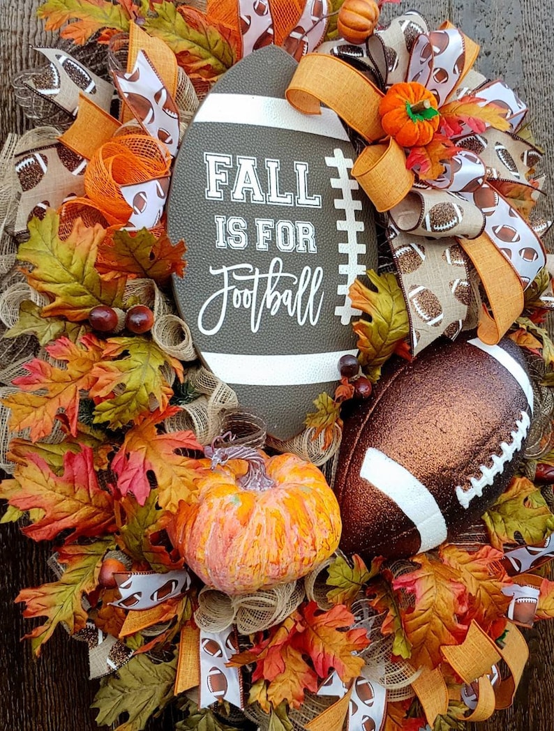 May include: A fall-themed wreath with a brown football-shaped sign that says "Fall is for Football". The wreath is decorated with orange and brown ribbons, fall leaves, and a small orange pumpkin.