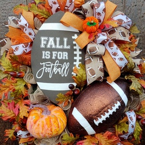 May include: A fall-themed door wreath with a brown football, orange pumpkins, and a sign that reads "Fall is for Football". The wreath is decorated with brown, orange, and white ribbons.