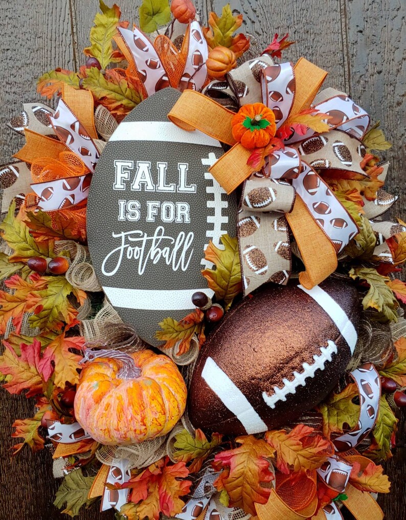 May include: A fall-themed door wreath with a brown football, orange pumpkins, and a sign that reads "Fall is for Football". The wreath is decorated with brown, orange, and white ribbons.