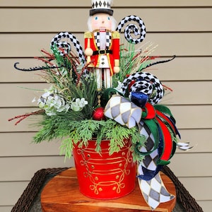 May include: A festive Christmas centerpiece featuring a wooden nutcracker soldier in a red coat and tall hat. The arrangement includes greenery, red berries, and decorative ribbons in a red bucket with gold accents, set on a wooden base.