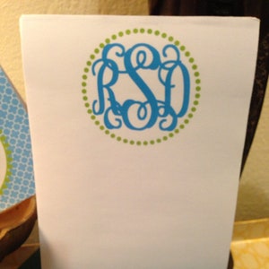 May include: White notepad with a teal monogram in a green dotted circle. A matching pen with a teal and white pattern and monogram rests below the notepad on a green surface. Stationery.