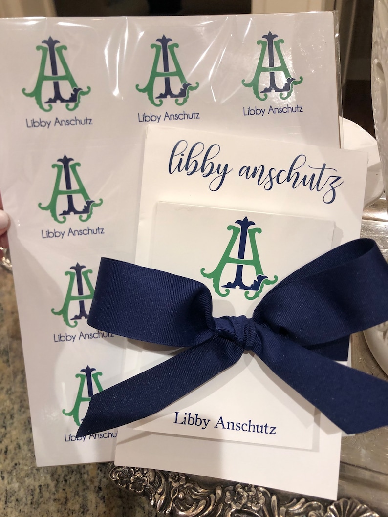 May include: A stationery set with a monogram design featuring the initials "A" and "I" in green and navy blue. The set includes notecards, stickers, and a navy blue ribbon. The text "Libby Anschutz" is printed on the items.