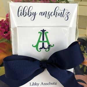 May include: A white stationery set featuring the name "Libby Anschutz" in navy script. The set includes a card with a green and navy monogram and a navy blue bow.