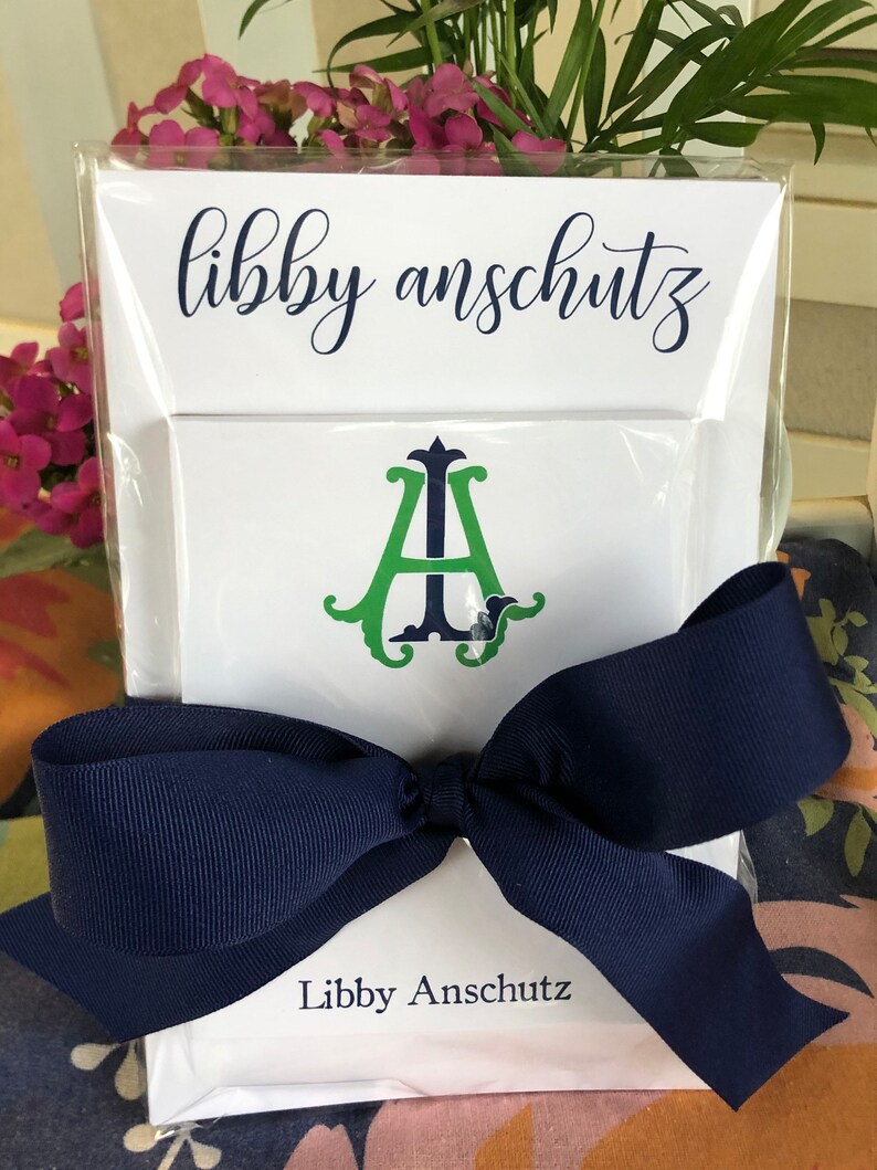 May include: A white stationery set featuring the name "Libby Anschutz" in navy script. The set includes a card with a green and navy monogram and a navy blue bow.