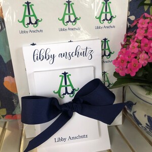 May include: A white stationery set tied with a navy blue bow. The set includes notepads and cards printed with the name "Libby Anschutz" in navy script. The stationery features a green and navy monogram design.