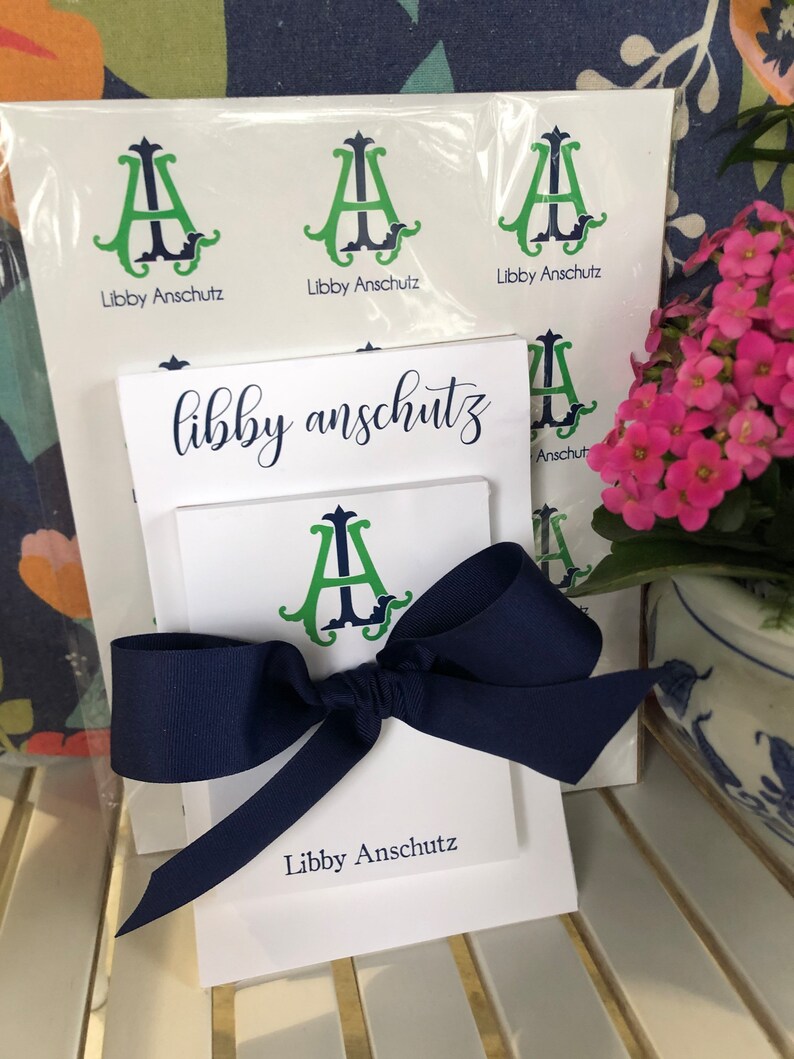 May include: A white stationery set tied with a navy blue bow. The set includes notepads and cards printed with the name "Libby Anschutz" in navy script. The stationery features a green and navy monogram design.