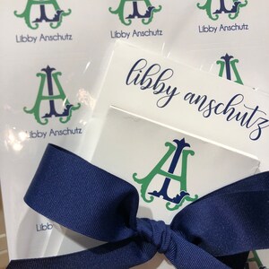 May include: White stationery with a navy blue ribbon. The stationery features a green and navy monogram with the name "Libby Anschutz" printed below. Several sheets are visible, some with the monogram and name, and others with just the name.