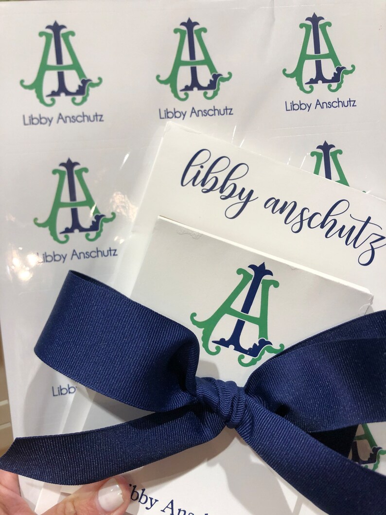 May include: White stationery with a navy blue ribbon. The stationery features a green and navy monogram with the name "Libby Anschutz" printed below. Several sheets are visible, some with the monogram and name, and others with just the name.