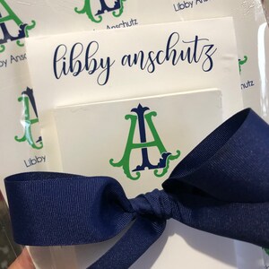 May include: White stationery featuring the name "libby anschutz" in navy script and a green and navy monogram "A" design. A navy blue ribbon is tied around the stationery.