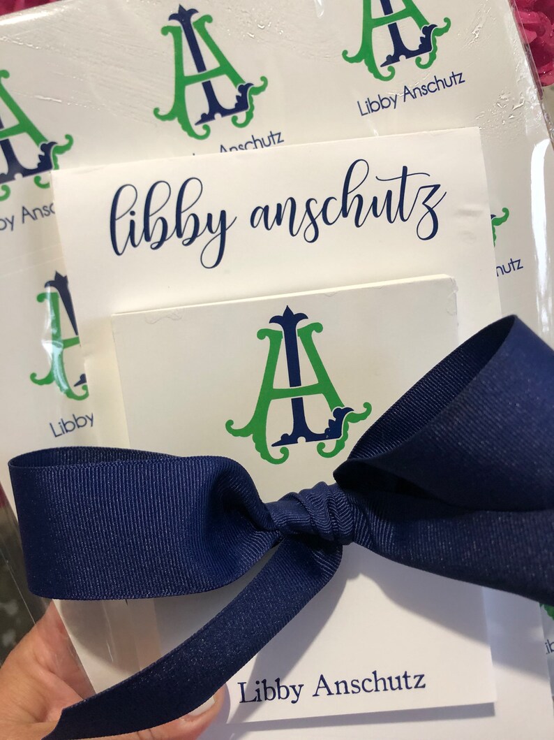 May include: White stationery featuring the name "libby anschutz" in navy script and a green and navy monogram "A" design. A navy blue ribbon is tied around the stationery.
