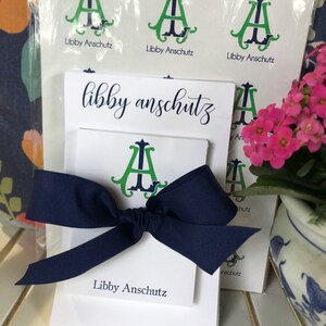 May include: A white stationery set with a navy blue bow. The set includes stickers and notecards featuring a green and navy monogram and the name "Libby Anschutz". A bundle of stickers is visible.