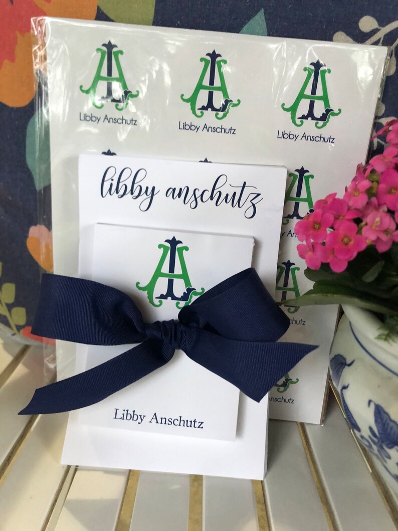 May include: A white stationery set with a navy blue bow. The set includes stickers and notecards featuring a green and navy monogram and the name "Libby Anschutz". A bundle of stickers is visible.