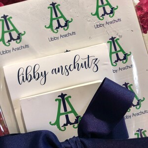 May include: A stationery set with the name "Libby Anschutz" in a handwritten font. The set includes stickers and cards with a green and navy blue monogram "A" design. A navy blue ribbon is tied around the set, with pink flowers in the background.