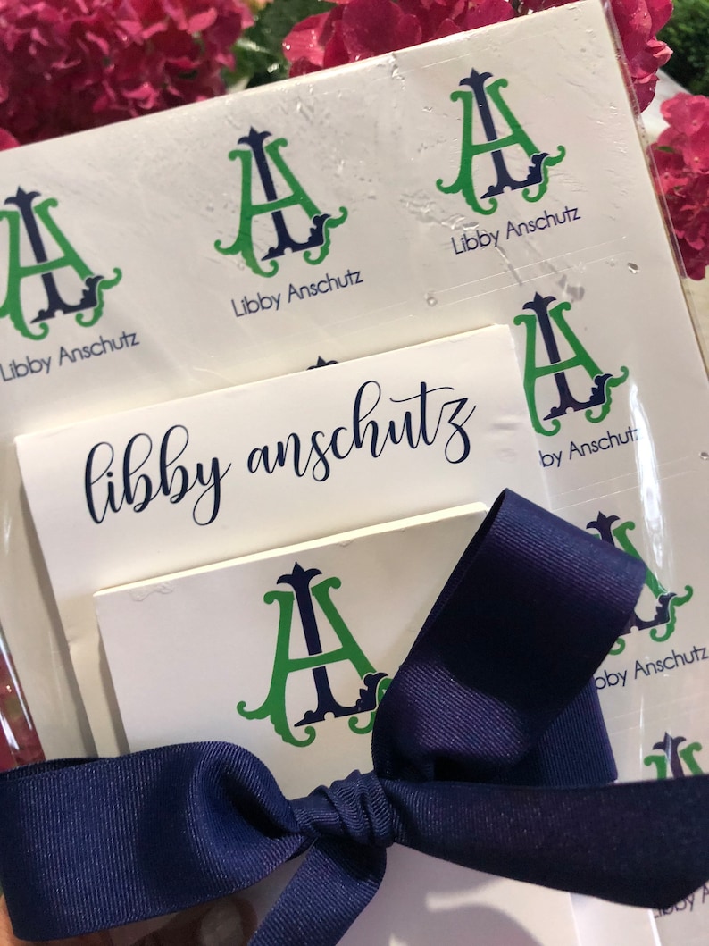 May include: A stationery set with the name "Libby Anschutz" in a handwritten font. The set includes stickers and cards with a green and navy blue monogram "A" design. A navy blue ribbon is tied around the set, with pink flowers in the background.