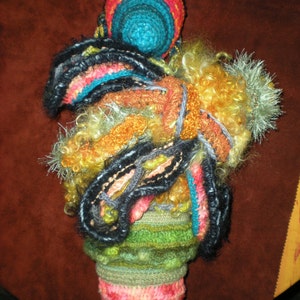 May include: A colorful crocheted doll with a round head and a long, flowing body. The doll is made with a variety of yarns in shades of pink, red, orange, yellow, green, blue, and black. The doll's head is topped with a crocheted hat.