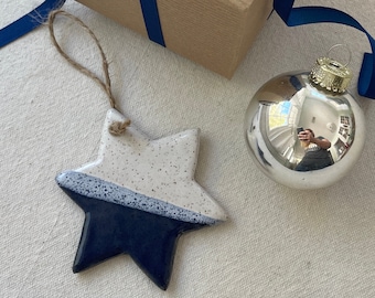 May include: A blue and white ceramic star ornament with a speckled glaze. The ornament is hanging from a twine string and is next to a silver glass ornament and a brown gift box with a blue ribbon.