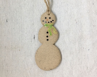 May include: A ceramic snowman ornament with a jute string for hanging. The snowman has a light brown speckled body, black dot eyes and buttons, and a green scarf. A festive holiday decoration.