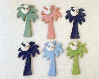 May include: Six ceramic palm tree ornaments in various colors, including green, pink, blue, and navy. Each palm tree has a white crescent moon on top.