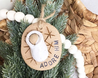 May include: A brown clay ornament with a white baby Jesus figurine and the text "Adore Him" in black lettering. The ornament is decorated with white snowflake designs.