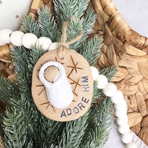 May include: A brown clay ornament with a white baby Jesus figurine and the text "Adore Him" in black lettering. The ornament is decorated with white snowflake designs.