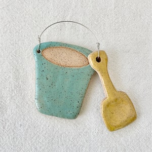 May include: A ceramic wall hanging featuring a teal blue bucket and a yellow shovel. The bucket has a beige rim and a speckled texture. The shovel has a speckled yellow glaze. The two pieces are connected by a silver wire.