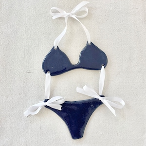 May include: A navy blue ceramic bikini with white ribbon ties. The bikini is a decorative piece and is not meant to be worn.