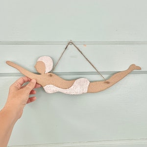 May include: A ceramic wall hanging depicting a swimmer in a vintage style. The swimmer is light brown with a white swimsuit and swim cap. The piece is suspended by a natural twine string.