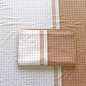 May include: A brown and white checkered fabric with a brown and white striped border. The fabric is folded in half and the striped border is visible on both sides.