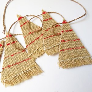 May include: A garland featuring multiple triangular burlap pennants suspended on a thin brown twine. Each tan burlap triangle is decorated with horizontal red stitched lines and finished with a frayed, textured bottom edge with gold thread.