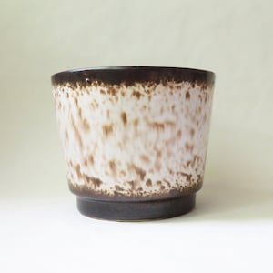 May include: A brown and white ceramic planter with a speckled glaze. The planter has a wide, rounded base and a slightly flared rim.