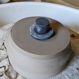 Może przedstawiać: Zbliżenie na wyrób ceramiczny z czarną gumową podstawą na kole garncarskim. Wyrób ceramiczny jest jasnobrązowy i ma gładką, lekko fakturowaną powierzchnię.
