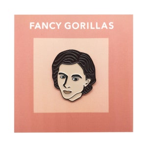 Puede incluir: Un pin de esmalte blanco y negro con el retrato de una persona con cabello castaño. El pin tiene un fondo blanco y el texto "FANCY GORILLAS" está impreso encima de la imagen.